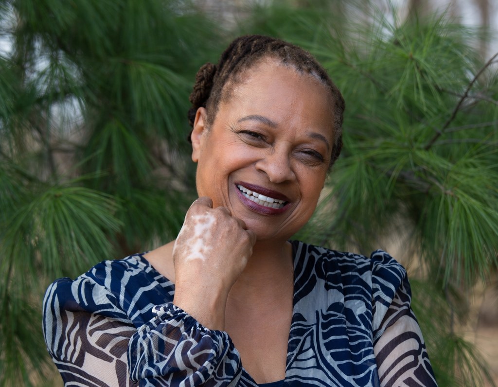 Ruth‑Ann Thompson smiling, hand resting at her chin, among pine trees