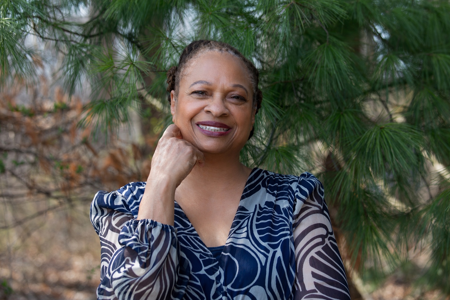 Ruth‑Ann Thompson outdoors among pine branches, smiling toward the camera